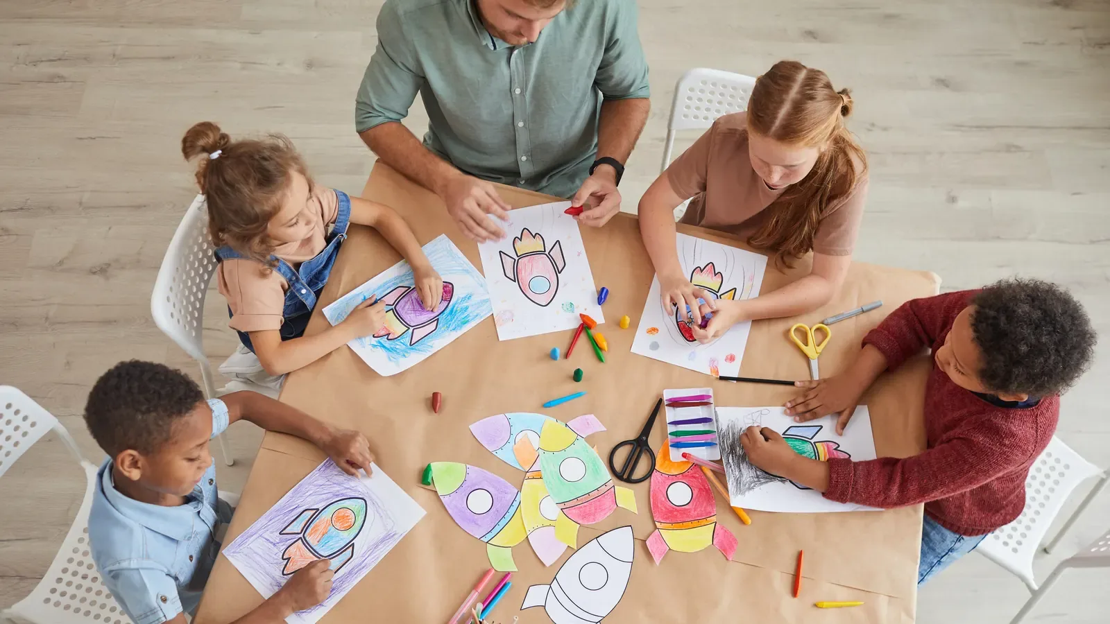 Four young children and two adults around a table from above, drawing rockets with crayons and felt-tip pens