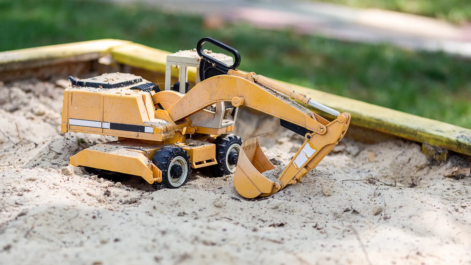 Yellow toy excavator digging in a wooden backyard sandbox