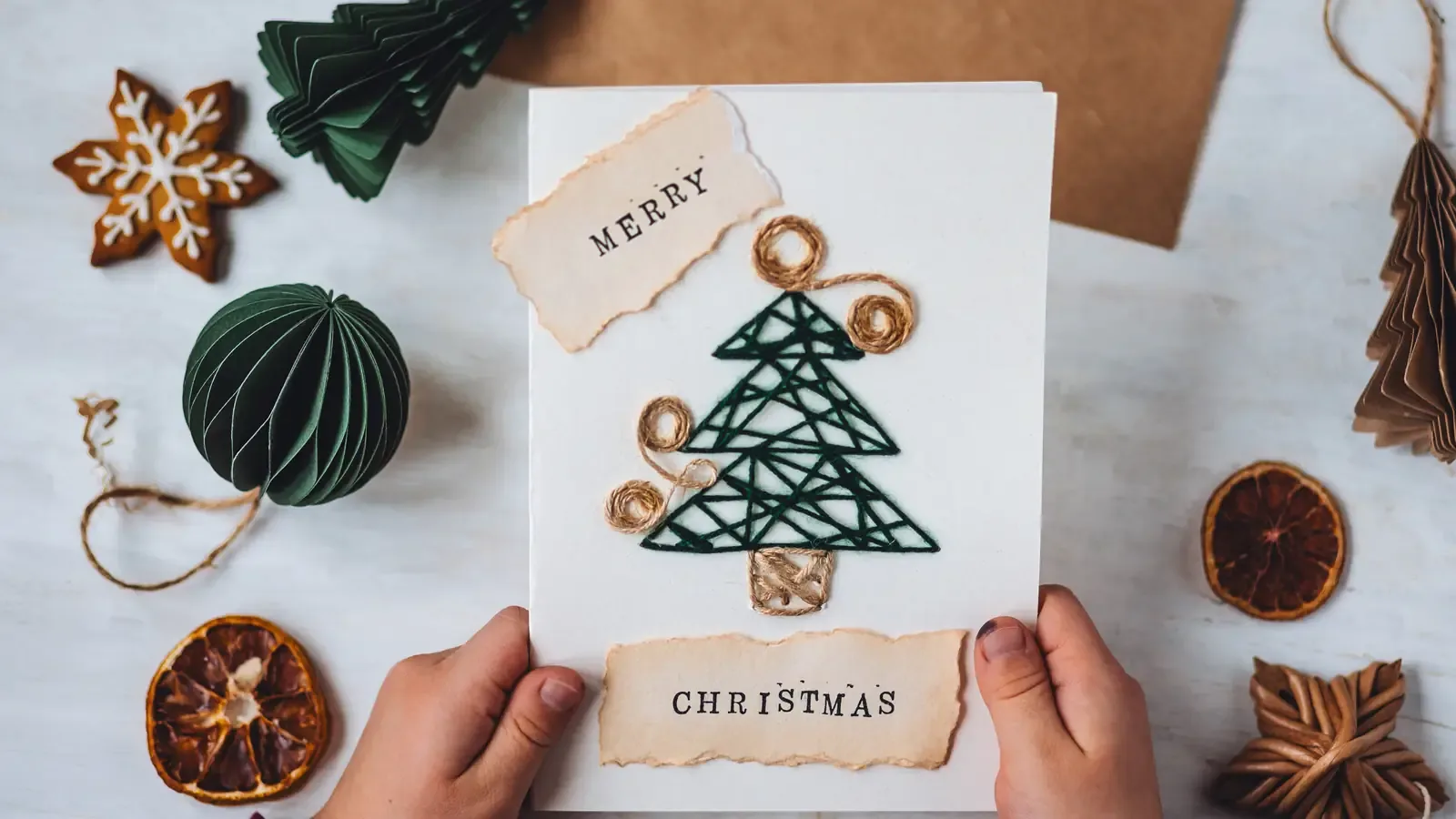 A child's hands holding a handmade Christmas card with a green string tree, surrounded by paper decorations and dried orange slices