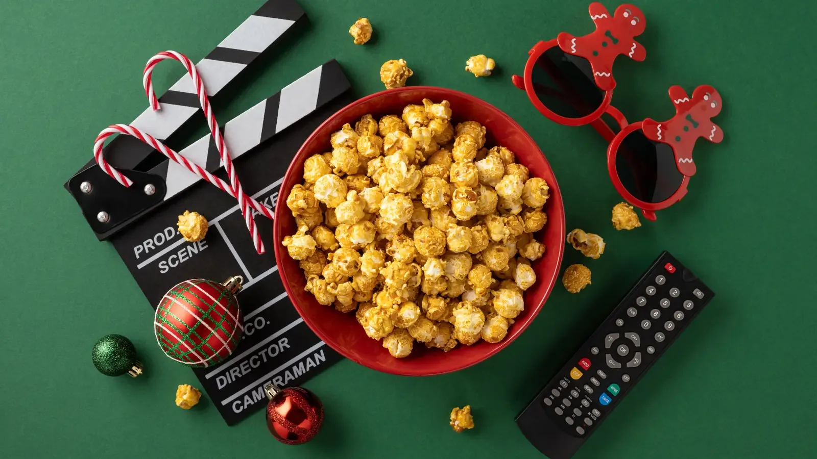 A red bowl of popcorn on a green background surrounded by a movie clapperboard, candy canes, Christmas ornaments and a TV remote