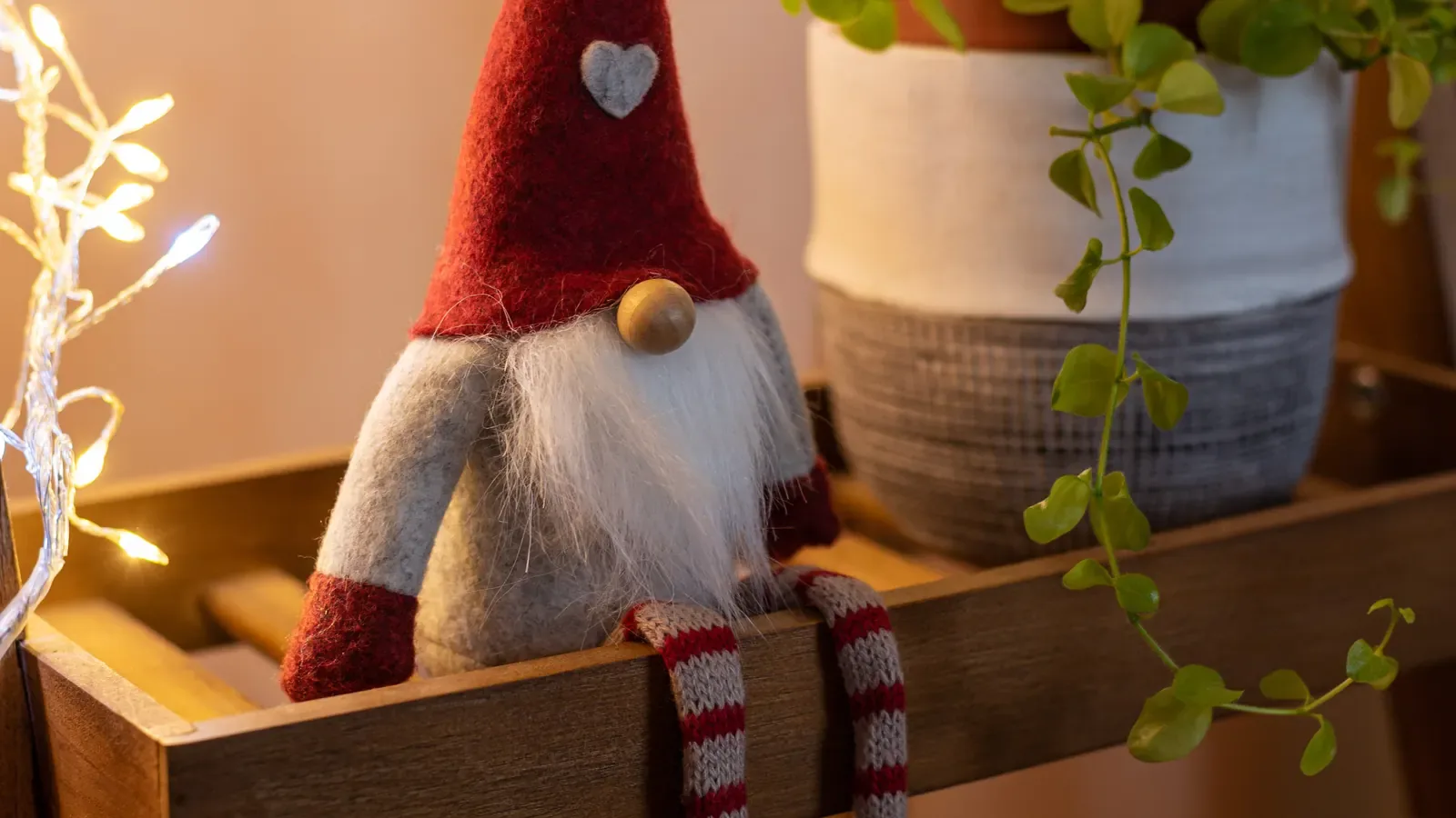 A red-hatted Christmas elf figurine sitting on a wooden shelf beside a green plant, lit by warm string lights