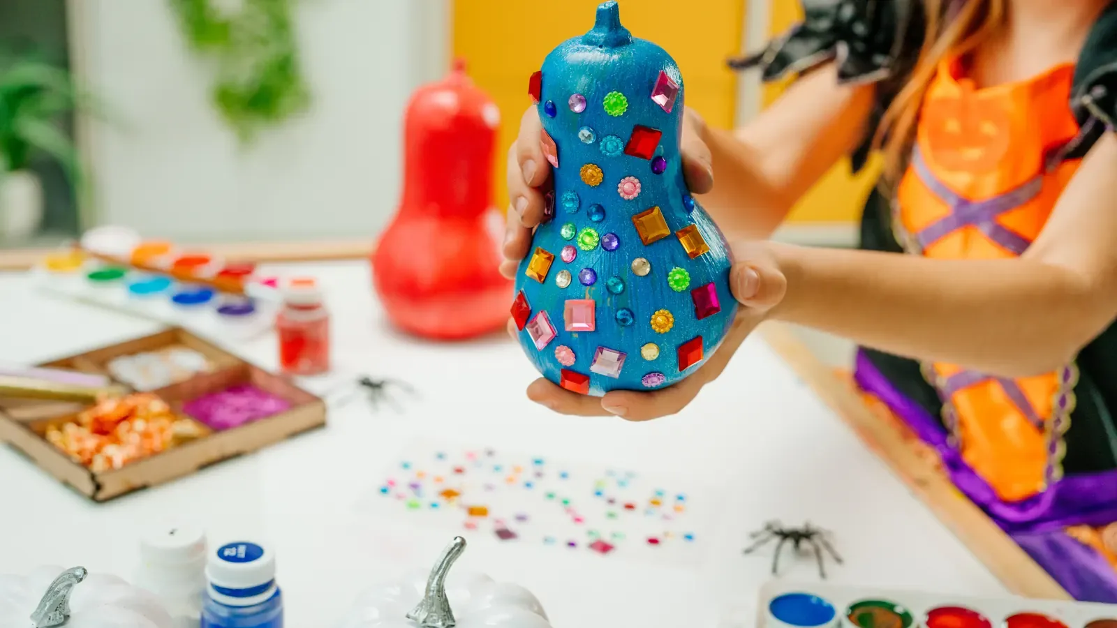 A child holding up a blue-painted pumpkin covered in colorful stickers, with paints and small white pumpkins on the table