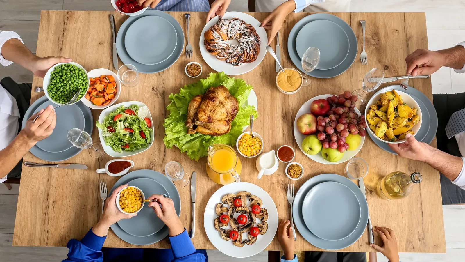 A Thanksgiving dinner table seen from above, with a roast turkey, salad, corn and side dishes, and family hands reaching for plates