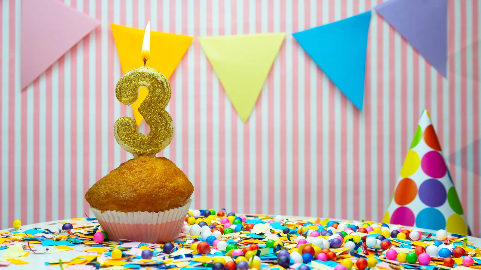 A gold glitter number 3 candle burning on a cupcake, with paper bunting, a party hat and scattered confetti