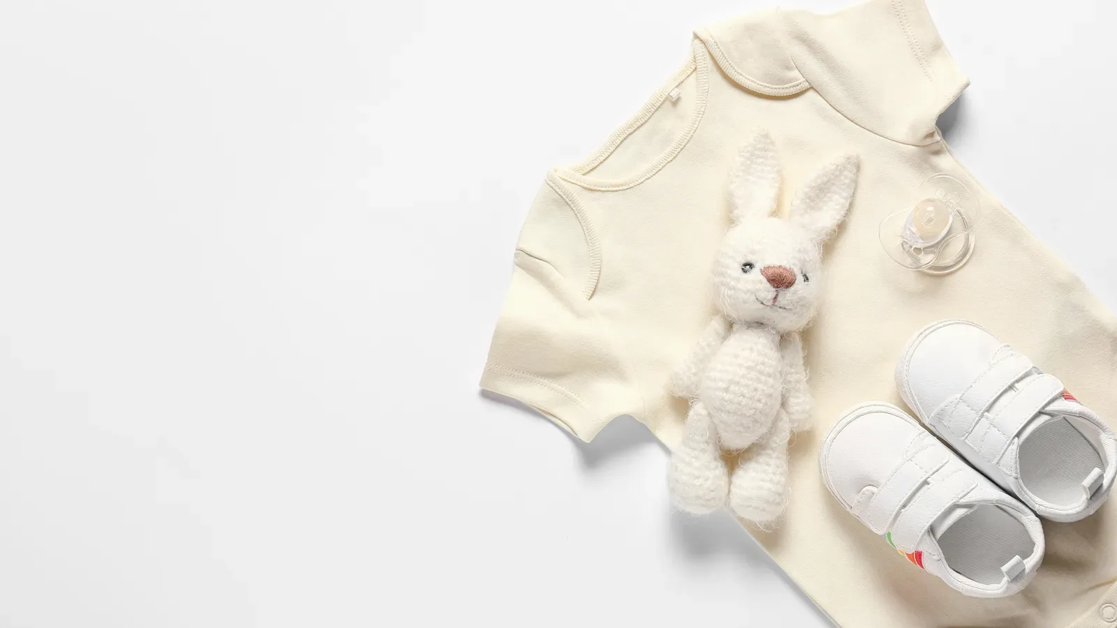 Baby clothes, shoes, a soft toy and a pacifier laid out on a white background