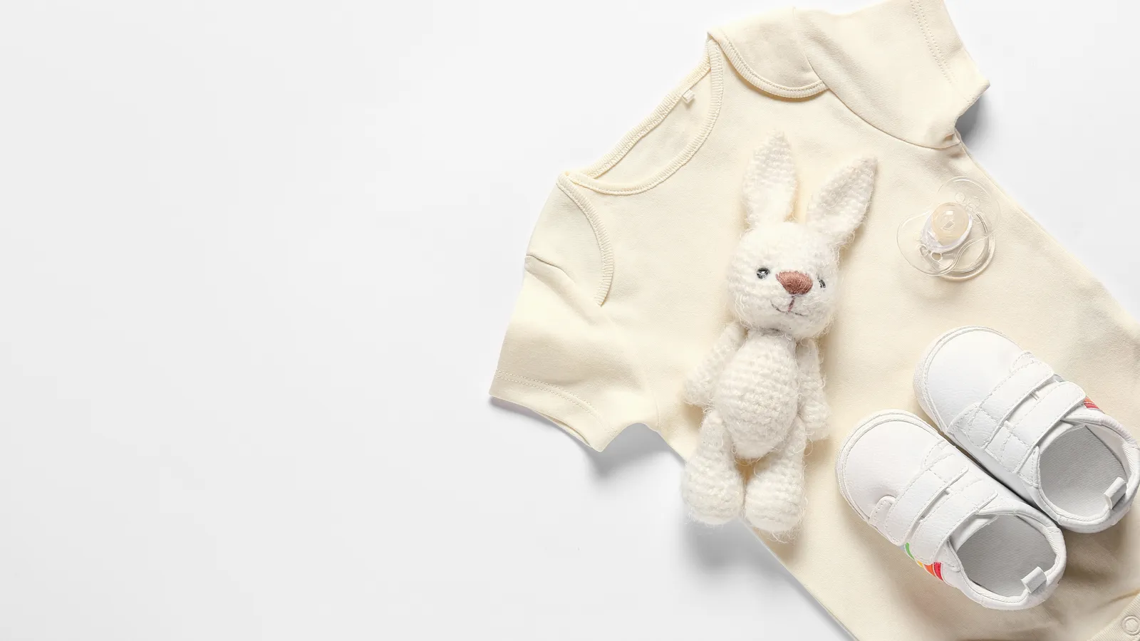 Baby clothes, shoes, a soft toy and a pacifier laid out on a white background