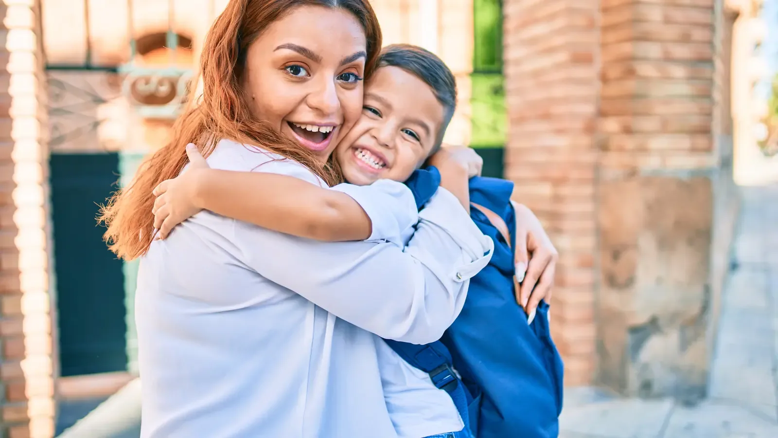 A mother and her young son hugging tightly and laughing in front of a brick wall