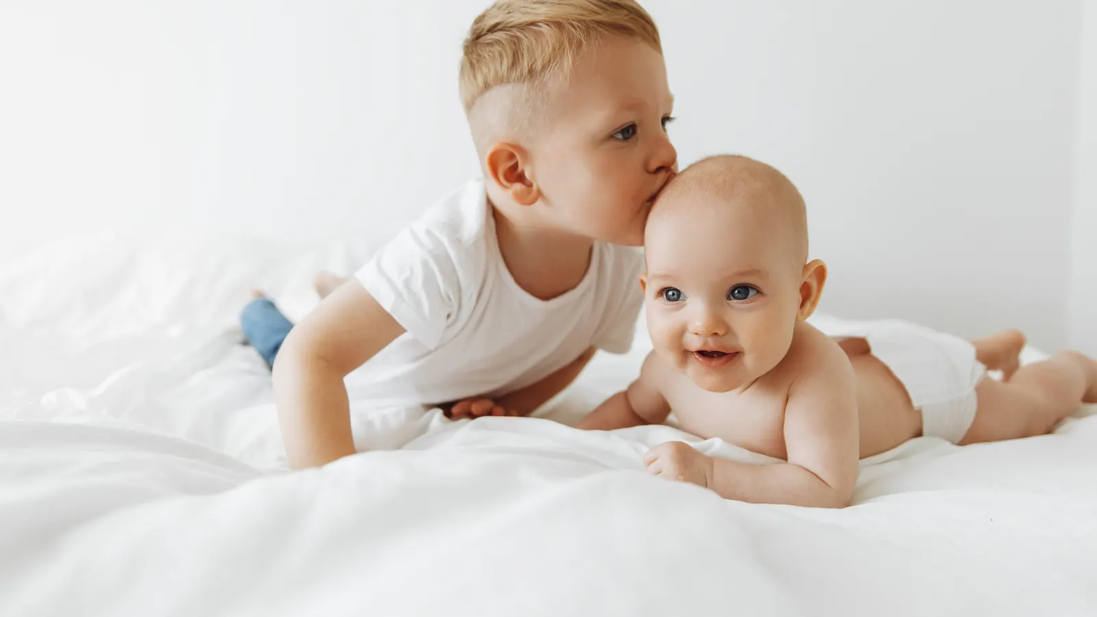 A young boy leaning over to kiss the head of a smiling baby lying on a white bed