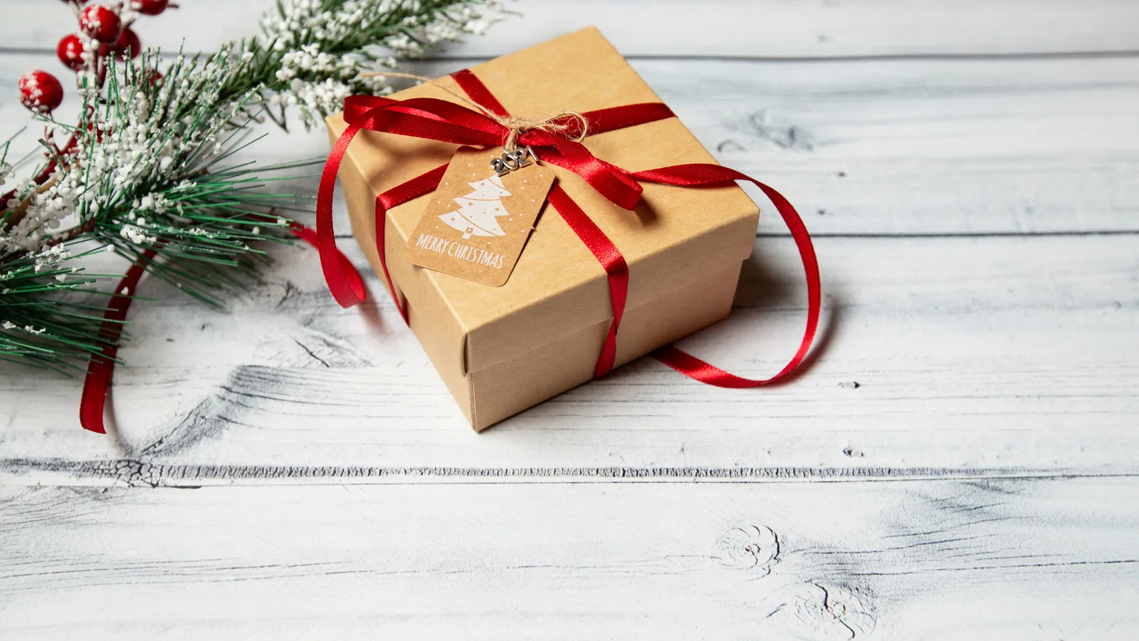 A single kraft gift box tied with red ribbon and a Merry Christmas gift tag, beside frosted pine and red berries on whitewashed wood