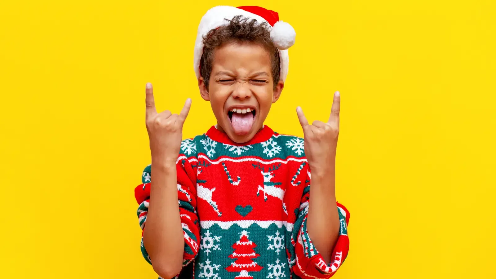 A ten-year-old boy in a Christmas sweater and Santa hat making a rock-and-roll hand gesture against a yellow background