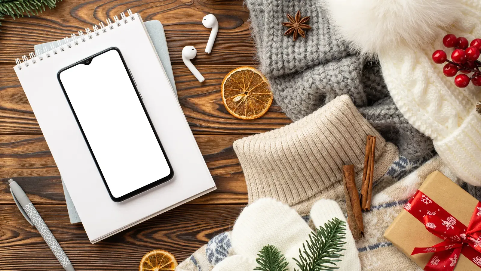 A flat lay of a smartphone, wireless earbuds, a notebook, a knit hat and a wrapped gift on a wooden desk
