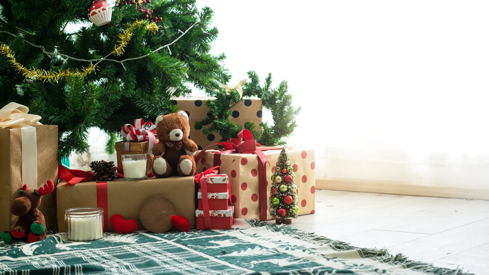 Wrapped gifts and a teddy bear piled under a decorated Christmas tree on a green checked rug