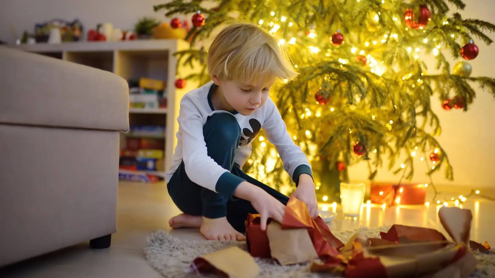A young boy kneeling on a rug unwrapping a present in front of a lit Christmas tree