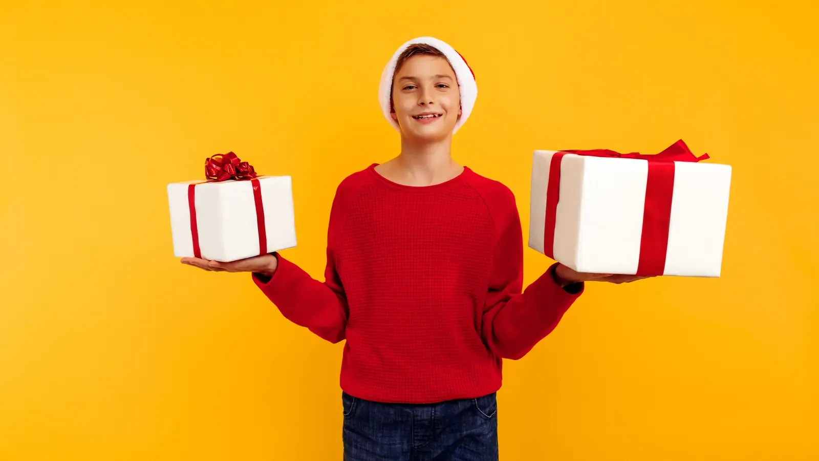 A young boy in a red sweatshirt holding two wrapped Christmas presents and wearing a Santa hat
