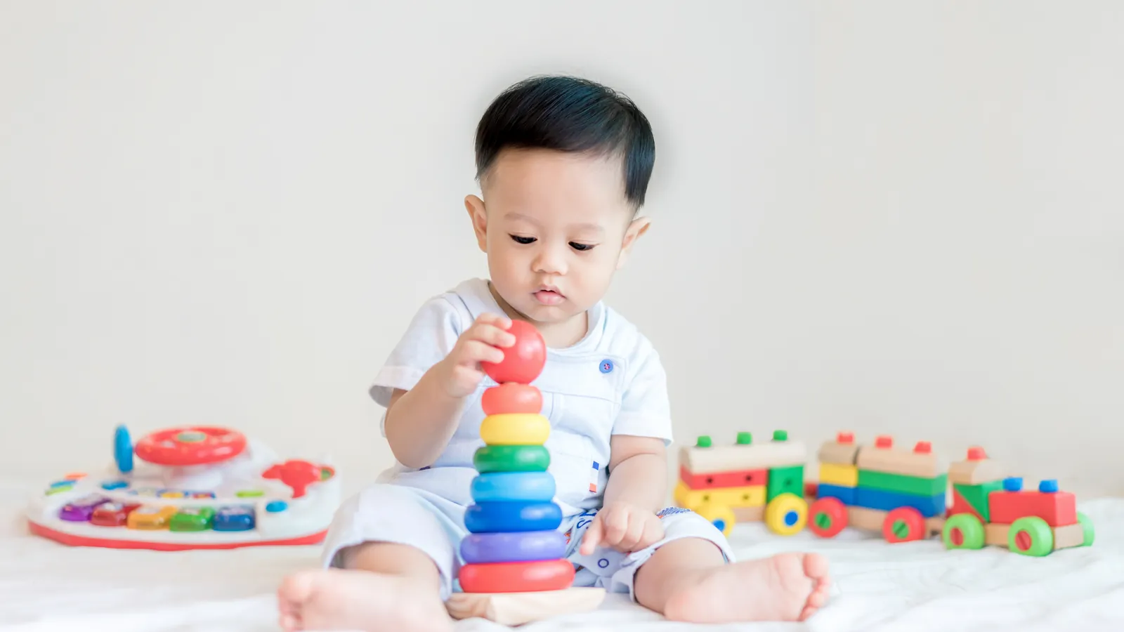 A baby boy sitting up on a white bed, reaching for the top ring of a stacking toy, with a wooden train and an activity toy beside him