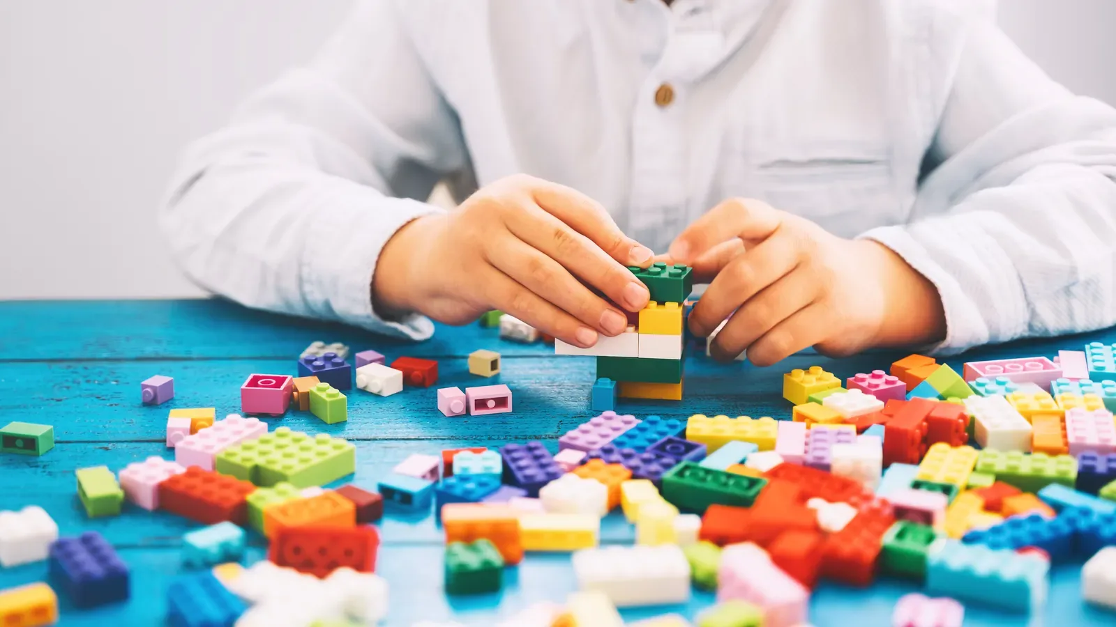 A child's hands joining two toy building bricks above a blue table scattered with colorful bricks