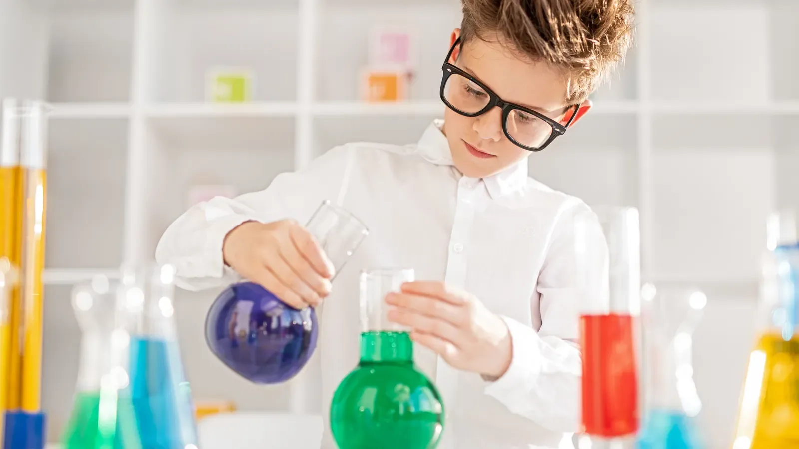 A boy in glasses holding a flask of blue liquid at a table of colorful science beakers