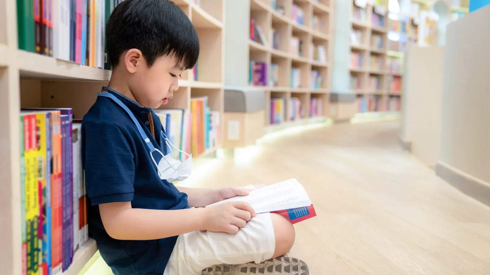 A boy sitting cross-legged on the floor of a library, reading an open book beside shelves of books