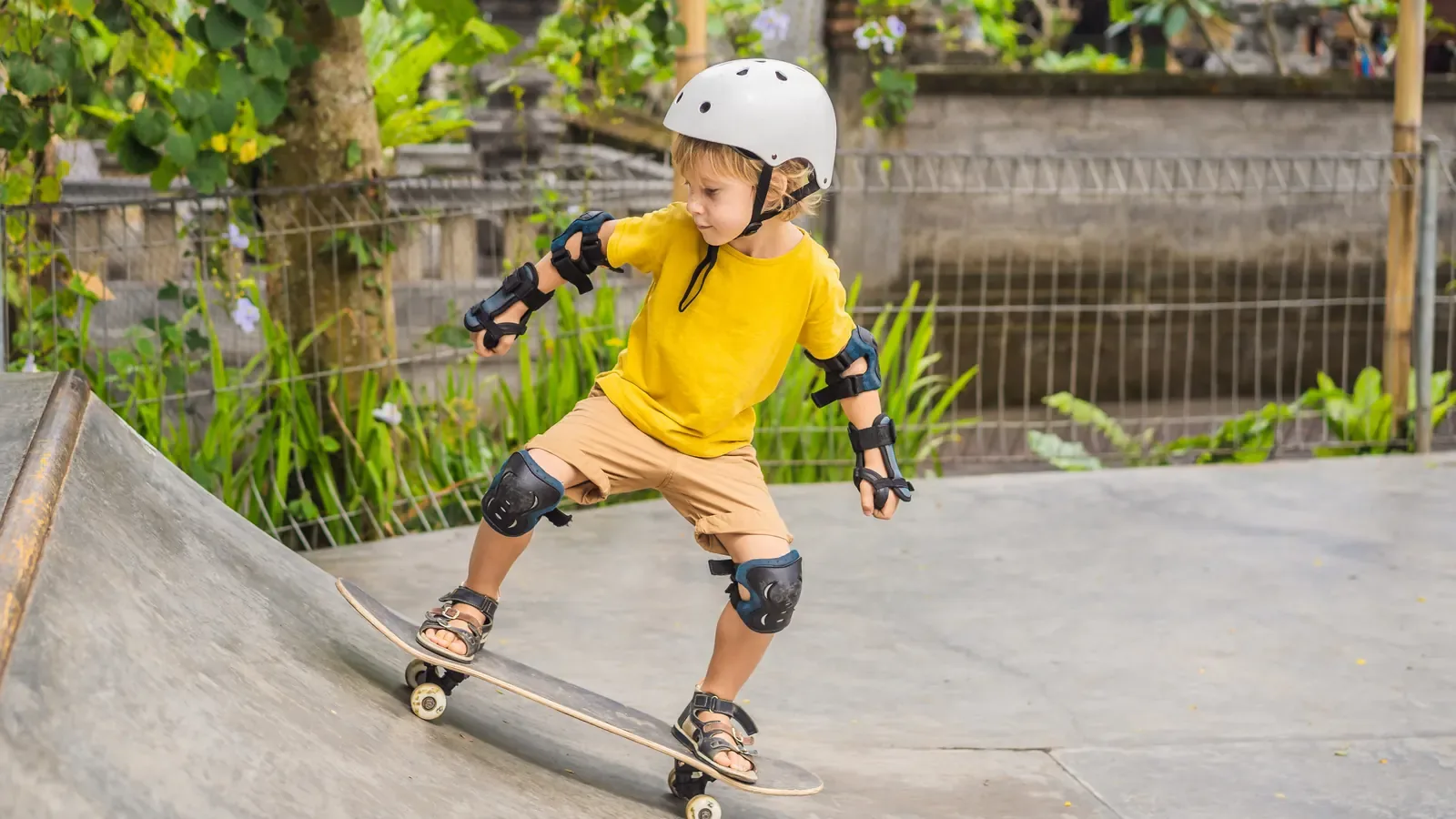 A boy in a helmet, yellow shirt and knee pads riding a skateboard down a ramp at a skate park