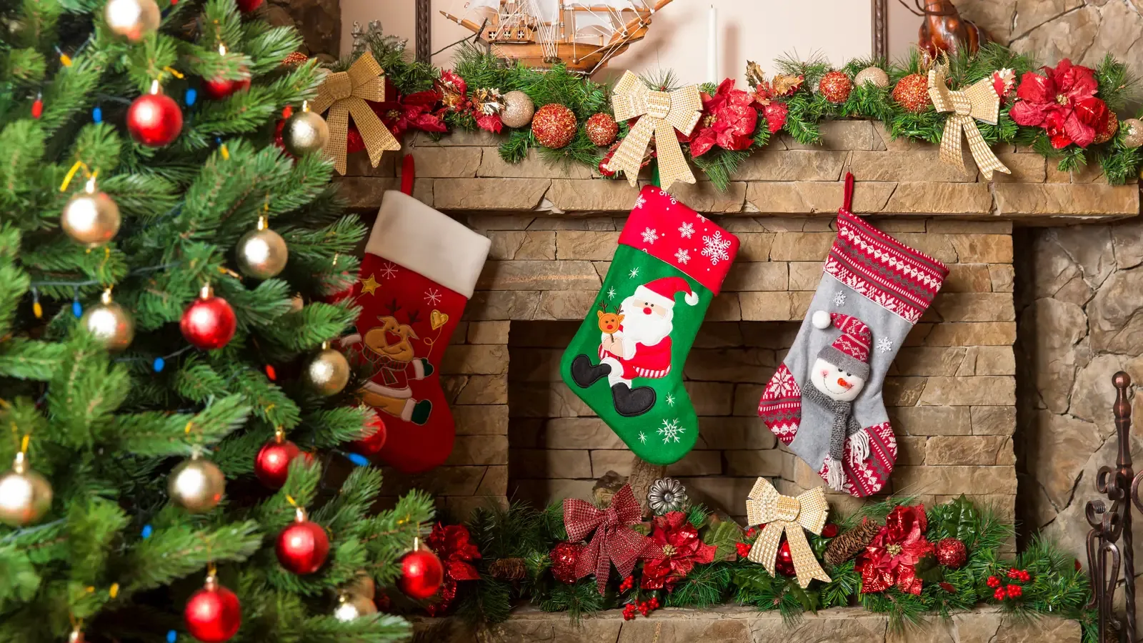 Three Christmas stockings hung on a stone fireplace decorated with garland, next to a lit Christmas tree
