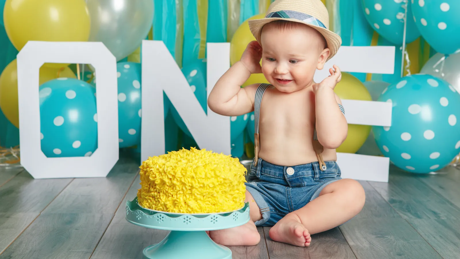 A baby boy in a straw hat and denim shorts sitting beside a yellow frosted cake, with large ONE letters and teal and yellow balloons behind him