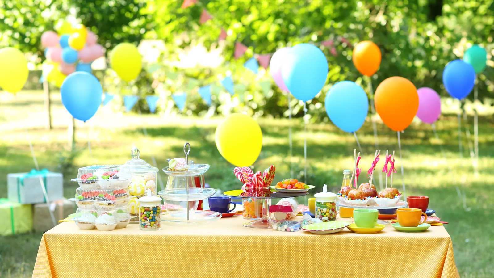 An outdoor party table with a yellow cloth, laid with cups, small plates of party food and a cake, under colorful balloons in a sunny garden