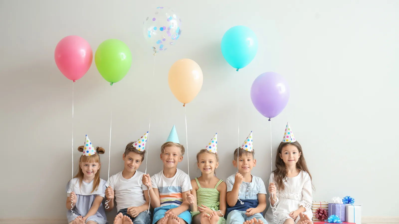 Five young children in paper party hats sitting in a row against a pale wall, with pastel balloons above them