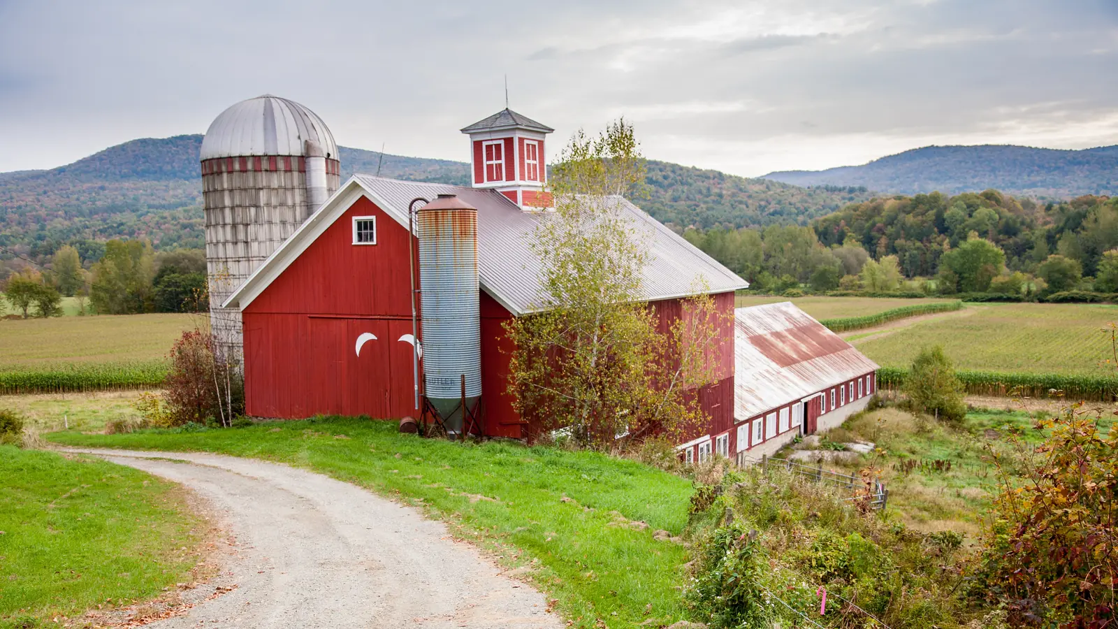 A classic red barn with a silo and white cupola beside a dirt track, with green fields and wooded hills behind