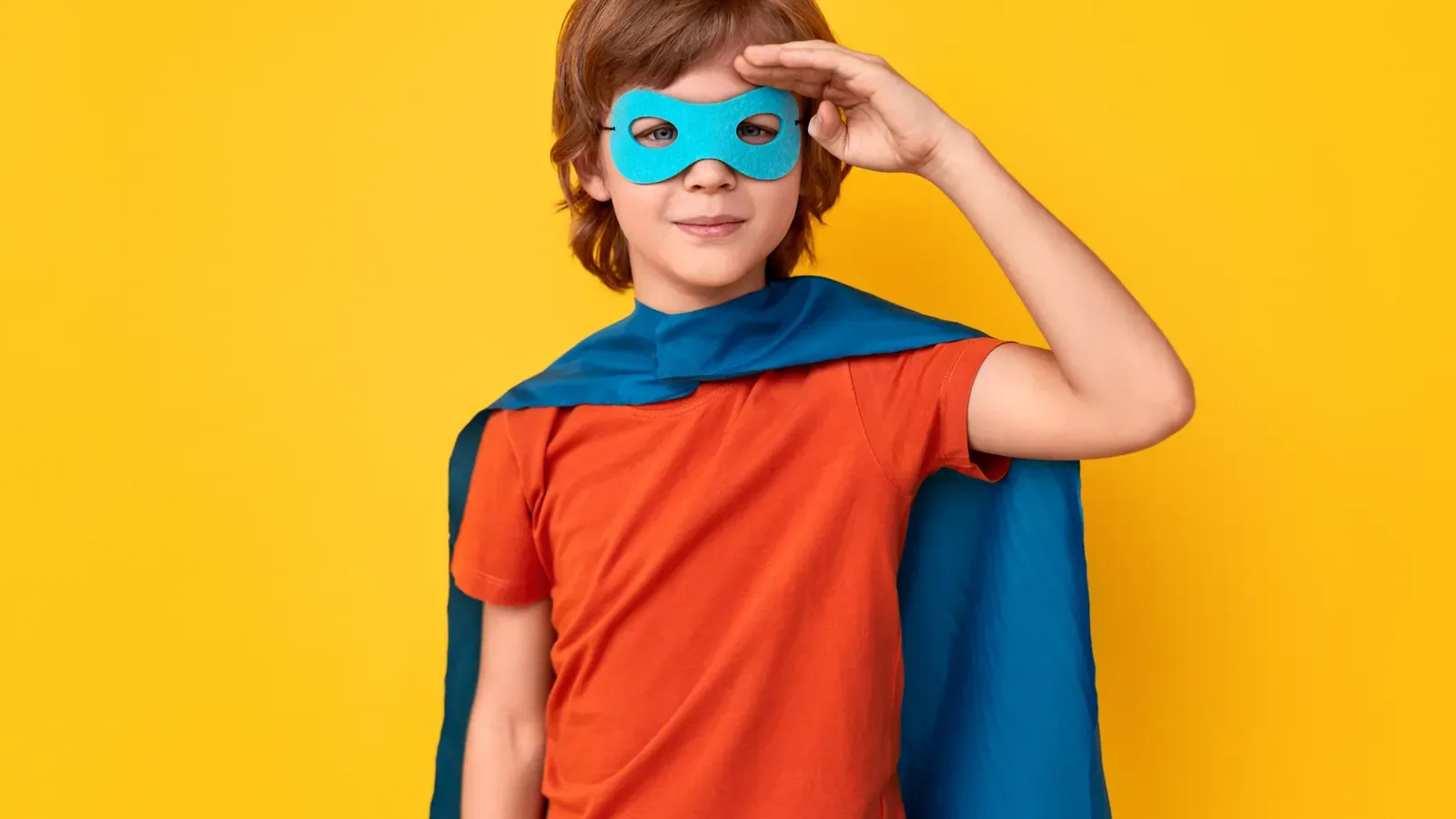 A child in a red shirt, blue cape and blue eye mask saluting against a bright yellow background